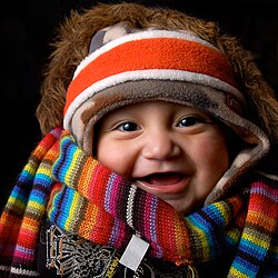 A baby wearing many items of winter clothing: headband, cap, fur-lined coat, scarf, and sweater