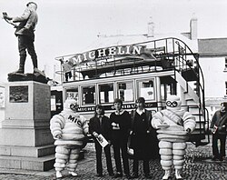 View of old-fashioned Michelin omnibus and two Michelin Men with bystanders behind Charles Rolls statue, Monmouth, Wales, c. 1965–1970