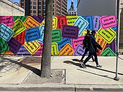 A vibrant mural of colorful "You Are Beautiful" stickers brightens a city street as pedestrians walk by.