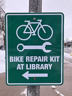 A picture of a green sign pointing to the left that reads "bike repair kit at library"