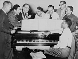 Goodman (third from left) with some of his former musicians, seated around piano left to right: Vernon Brown, George Auld, Gene Krupa, Clint Neagley, Ziggy Elman, Israel Crosby and Teddy Wilson (at piano); 1952