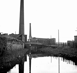 A black-and-white photograph of a canal taken from a boat on it. On the left-hand side is a small building with a massive chimney stretching above the top of the photo. To the right of the canal is a much bigger building, itself also with a massive chimney, all of which is in the photo. The right-hand building appears directly in front due to the curvature of the canal, and a bridge goes over the canal between the two buildings.