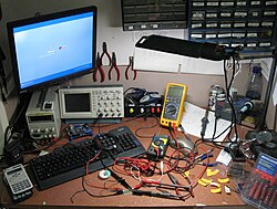 The workbench of a hobby electronics enthusiast with an oscilloscope, PC, two digital multimeters, soldering station, pocket calculator, small tools, a stabilized power supply unit and storage boxes on the wall for electronic components
