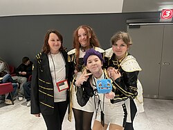 Group shot of four people wearing cosplay. The person on the left is dressed as Bobby. The other three are wearing Huntrix "Golden" outfits; one person is slightly crouched in front of the other two, holding a small blue tiger plush.