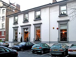 A number of cars are parked in the asphalt car-park of a two-storey white building. The building appears once to have been detached, but a two-storey extension is apparent, on the right side of the building. The ground floor contains three large sash windows, each with a small metal railing around the sill. The first floor contains four shorter sash windows. The decorative stonework around the windows and main entrance has been painted grey. Two chimney stacks are visible on the roof, at opposite ends. The staircase entrance to the building is bordered by metal railings. To the left of the image, a much larger brick building can be seen in the distance.