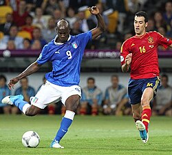 Spain holding midfielder Sergio Busquets (16, red) moves to block a shot from Italian striker (9, blue) Mario Balotelli.