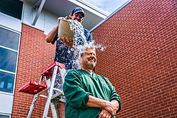 A person stands still and winces as a bucket full of ice and water is poured onto their head by another.
