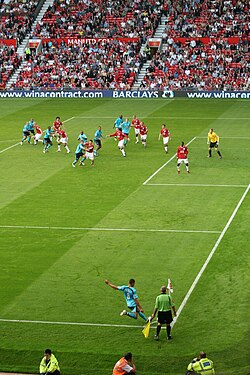 Jermaine Jenas (Tottenham Hotspur) taking a corner kick against Manchester United. The Tottenham players (blue) attempt to move into position to direct the corner towards the goal, while the United players (red, with goalkeeper in yellow) attempt to defend. The assistant referee (green, with flag) ensures that the corner is taken from inside the quarter-circle area.