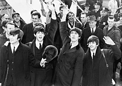 Black and white picture of the Beatles waving in front of a crowd with an set of aeroplane steps in the background