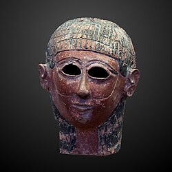 An ancient mask. The individual has brown skin, and black hair. There are holes at the eyes. Lines go down the face that are interpreted as a beard.