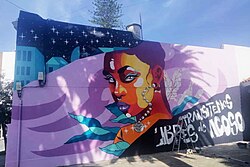 Outdoor mural in progress, with a starry night sky in the background and a Black woman with makeup and jewelry looking back at the viewer. Below, the words "TRANSITEMOS LIBRES de ACOSO" are painted.