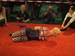 A female rigger stands by while a female model she has tied up tries to free herself
