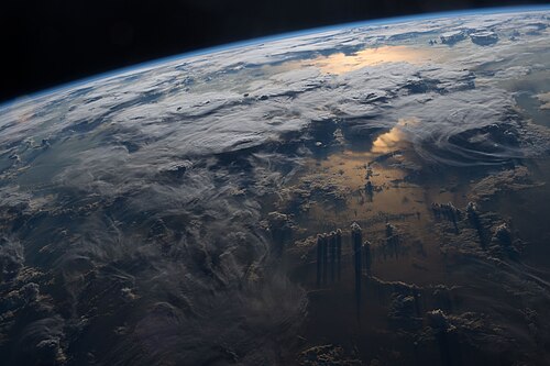 A picture of Earth's troposphere with its different cloud types of low to high altitudes casting shadows. Sunlight is reflected off the ocean, after it was filtered into a reddish light by passing through much of the troposphere at sunset. The above lying stratosphere can be seen at the horizon as a band of its characteristic glow of blue scattered sunlight.