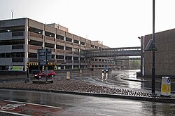 High level shot of multi lane road lower down stretching into distance with two glass sided pedestrian walkways over linking car park to left and shopping centre to right