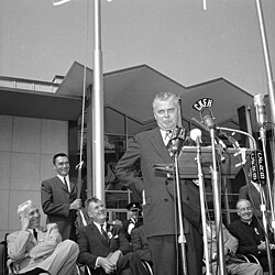 A man in suit and tie stands with arms on hips behind a podium with media microphones attached.