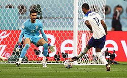 Marcus Rashford (no. 11, in white) is 1 on 1 against Hossein Hosseini (no. 24, in blue) and is attempting to score. The goalkeeper will try to stop the forward from scoring a goal by preventing the ball from passing the goal line.