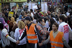 A crowd of people marches with banners. At the edge of the crowd, men in orange vests hold hands and look out from the crowd to form a border.