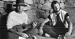 Two cross-legged men sit against a blocky wall. The one on the left holds a string instrument
