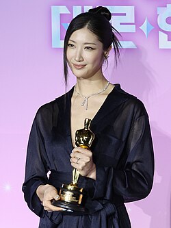 Ejae from the waist up holding her Oscar trophy with both hands. She is looking slightly left of center.
