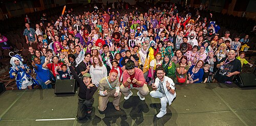 Group shot taken from the stage, facing the audience. The celebrities are crouched down with their backs to the audience while the audience is mostly standing to pose for the photo. Many people in the audience are wearing cosplay.