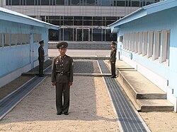 North Korean soldiers standing guard at the JSA between the blue buildings. View from the north. To the rear, the ground floor of Freedom House, in South Korea. The low-level concrete ledge running between the two soldiers is the border mark in the security area.