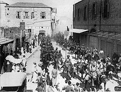 In a black and white photograph, a large group of turbaned men on horseback ride through a dusty, sunlit street and into the distance, obscured by dust. A crown of civilians watch the men pass. A large, brick-built, two-storey building is on the left, and a similar structure is on the right.