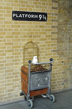 A sign reading "Platform .mw-parser-output .frac{white-space:nowrap}.mw-parser-output .frac .num,.mw-parser-output .frac .den{font-size:80%;line-height:0;vertical-align:super}.mw-parser-output .frac .den{vertical-align:sub}.mw-parser-output .sr-only{border:0;clip:rect(0,0,0,0);clip-path:polygon(0px 0px,0px 0px,0px 0px);height:1px;margin:-1px;overflow:hidden;padding:0;position:absolute;width:1px}9+3⁄4" with half of a luggage trolley installed beneath, at the interior of King's Cross railway station.